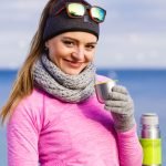 Woman athlete fit girl wearing warm sporty clothes outdoor in cold weather drinking hot tea from vacuum flask thermos warming up. Sports activities hiking in winter or autumn time
